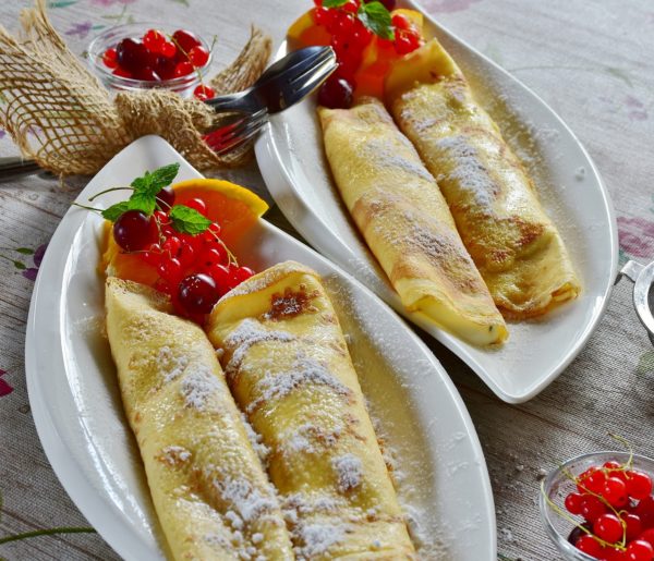 Crêpes saupoudrées de sucre glace, fourrées de nougat et accompagnées de fruits rouges et de tranches d'orange, servies dans une assiette blanche sur une nappe à motifs.